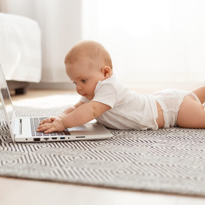 Little curious child studying modern technology while lying on floor on tummy against window, toddler using laptop at home, infant wearing white t shirt.